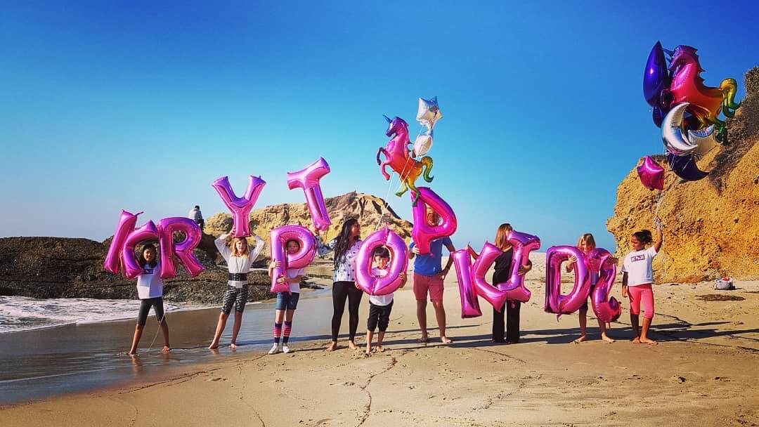 Krypto for Kids — the iconic beach photo: children and families spell out KRYPTO KIDS with giant pink balloon letters on Laguna Beach, California, November 2018. Unicorn balloons and rainbow decorations fill the sky.