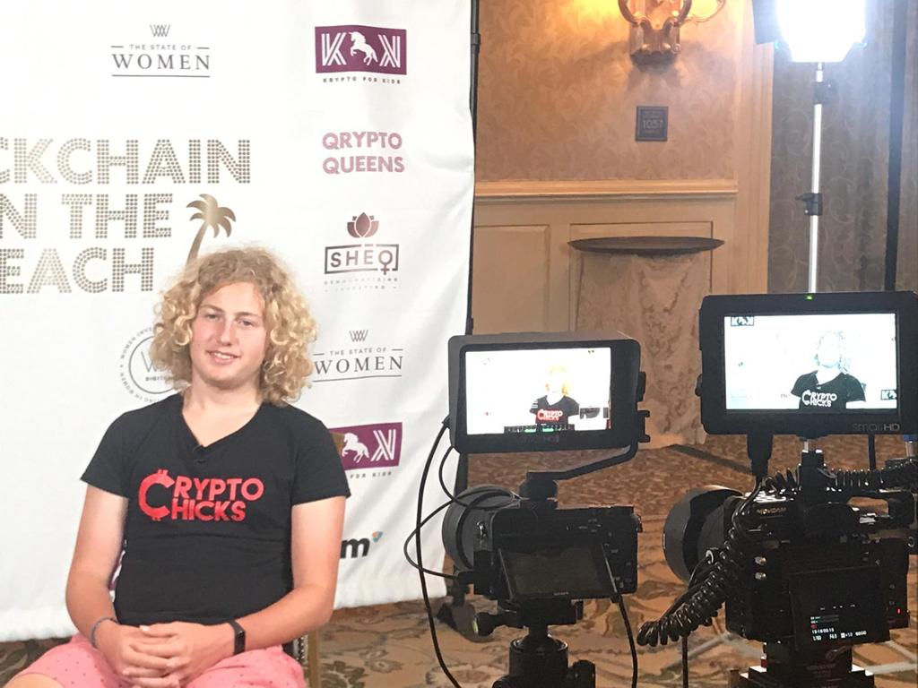 Blockchain on the Beach media interview — Arya Bhardwaj (in CryptoChicks t-shirt) being filmed in front of the Blockchain on the Beach step-and-repeat banner featuring Krypto for Kids, Qrypto Queens, SHEQONOMI, The State of Women, and NEM logos, Laguna Beach 2018.