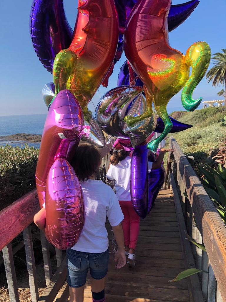 Krypto for Kids — children carrying rainbow unicorn and star balloons down the Laguna Beach boardwalk path toward the ocean, November 2018.